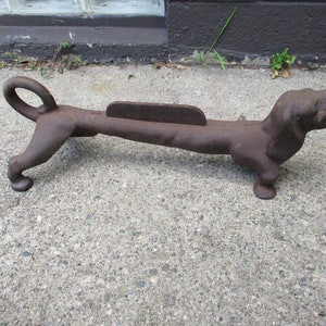 May include: A cast iron dachshund doorstop in a rusty brown color. The doorstop is lying on a gray concrete surface.