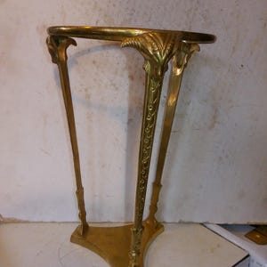 May include: A gold-colored, ornate plant stand with a round top and three legs. The legs are intricately designed with floral and leaf motifs. The stand is approximately 30 inches tall.