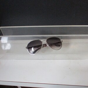 May include: Aviator sunglasses with a silver frame and gradient lenses rest inside a clear acrylic display shelf. The sunglasses have a classic design, suitable for various styles. The shelf is on a white surface.