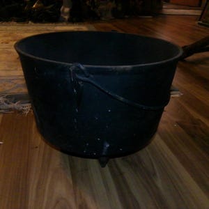 May include: A large, black cast iron cauldron with a single handle. The cauldron has a rough, textured surface and appears to be very old.