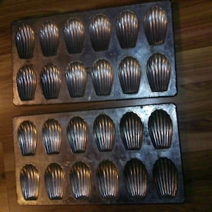 May include: Two rectangular metal baking pans, each featuring twelve shell-shaped indentations for making madeleines. The pans have a dark, weathered finish with visible signs of use and a metallic sheen across the molded cavities.