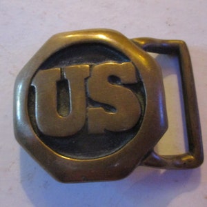 May include: A brass belt buckle with a raised octagonal design featuring the letters "US" in the center.