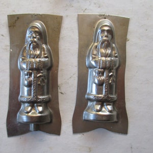 May include: Two vintage metal molds for making chocolate Santa Claus figures. The molds are silver in color and have a detailed design of Santa Claus holding a bag of gifts.