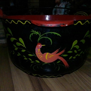 May include: A black, round, wide-mouthed basin with a red interior and rim. The exterior features a hand-painted red bird with yellow accents, surrounded by green leaf motifs and a yellow wavy line pattern along the top edge and base.