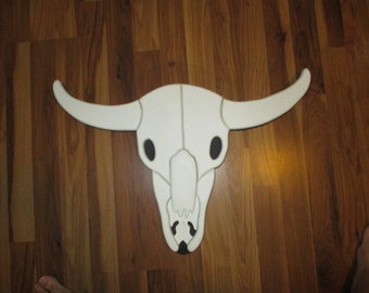 May include: White wooden bull skull with black eye sockets. The skull is mounted on a wood floor.