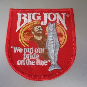 Puede incluir: Parche rojo bordado con un contorno blanco. El parche presenta a un hombre de dibujos animados sosteniendo un pez y el texto "BIG JON®" y "We put our pride on the line."