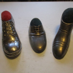 May include: Three metal shoe-shaped pincushions. The pincushions are in the shape of a dress shoe, a loafer, and a boot. The dress shoe has a red cushion, the loafer has a green cushion, and the boot has a green cushion.