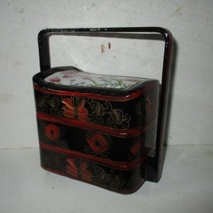 May include: A black, three-tiered, lacquered food carrier with a handle. The carrier has red accents and floral and geometric designs. The top tier features a white surface with a floral pattern. The handle is black.