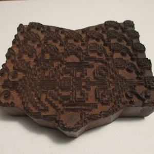 May include: A carved wooden printing block with a complex, repeating pattern. The block is a dark brown color and has a star-like shape. The intricate design suggests it could be used for textile printing or decorative purposes.