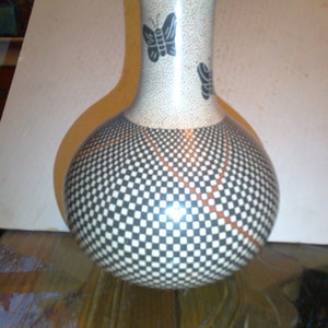 May include: A white ceramic vase with a black and white checkered pattern and red accents. The vase has a round body and a narrow neck with two black butterfly designs.