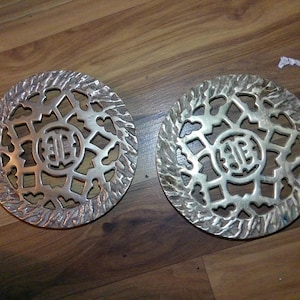 May include: Two round, silver-colored metal trivets with intricate openwork designs. Each trivet features a central emblem surrounded by a decorative border. The trivets are displayed on a wooden surface, showcasing their detailed craftsmanship.