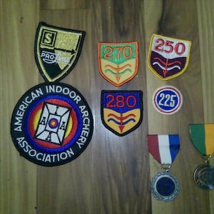 May include: A collection of archery patches and medals. The patches feature various designs, including the American Indoor Archery Association logo, scores, and the Pro Line logo. Medals with colored ribbons are also present.