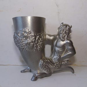 May include: A silver-colored goblet with a satyr figure handle. The goblet has a flared shape and is decorated with grape vines. The satyr is kneeling and has horns, a beard, and a muscular physique. The goblet is made of metal.