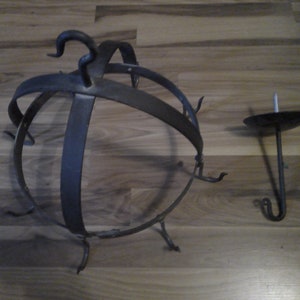 May include: A wrought iron candle holder and a separate candle cup. The candle holder is a spherical cage design with curved metal bars and decorative hooks. The candle cup has a small, round dish and a hook.
