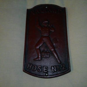May include: A dark brown, cast iron plaque with a relief of a firefighter in action. The plaque is rectangular with a curved top and the words "HOSE NO 2" are at the bottom. There are two holes for mounting.