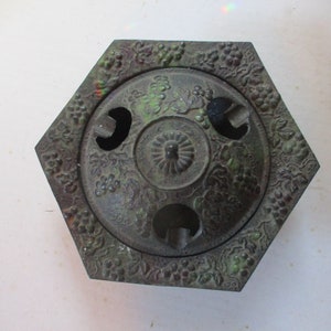 May include: A hexagonal cast iron ashtray with a green patina. The ashtray has three circular openings and is decorated with a grapevine design.