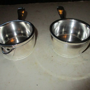 May include: Two small, silver-colored saucepans with handles. Each has a rounded body and a handle extending from the side. The rims are slightly darker. The saucepans are sitting on a light-colored surface.