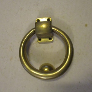 May include: A brass door knocker with a round ring and a ball-shaped knocker. The knocker is attached to a square plate with four screws.