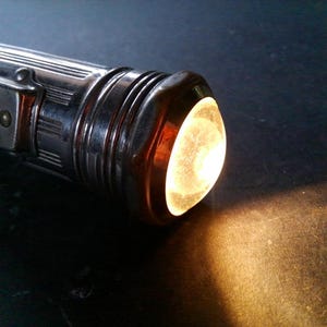 May include: A vintage, chrome-plated flashlight with a bright, warm light emanating from the lens. The flashlight has a ribbed design and a small switch on the side. The light casts a warm glow on the dark surface.