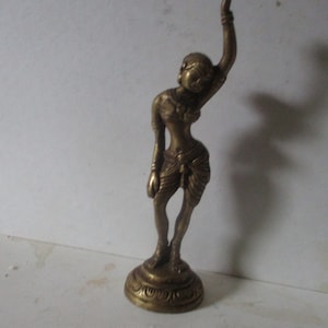 May include: A bronze-colored figurine of a dancing figure with one arm raised. The figure is wearing a draped garment and is standing on a circular base with a decorative pattern. The statue has a detailed face and is likely a representation of a deity or dancer.