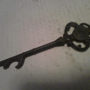 May include: A vintage-style metal key with a bottle opener attached to the end. The key has a decorative, ornate design with a circular opening in the center.