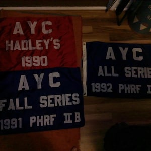 May include: Three rectangular flags with white grommets on the left side. One red flag reads AYC HADLEY'S 1990. Two blue flags read AYC FALL SERIES 1991 PHRF IIB and AYC ALL SERIES 1992 PHRF IIB. The text on all three flags is in white block lettering.