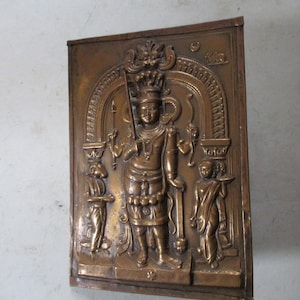 May include: A brass relief sculpture depicting a Hindu deity with a crown and ornate jewelry. The deity is standing in a doorway with two smaller figures on either side. The sculpture is mounted on a wooden plaque.