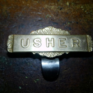 May include: A vintage brass "USHER" name badge with a decorative floral design. The rectangular nameplate is attached to a curved metal clip. The badge is set against a dark, textured background.