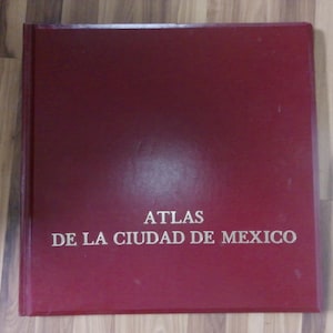 Puede incluir: Un atlas granate titulado "ATLAS DE LA CIUDAD DE MEXICO" en letras mayúsculas blancas. El libro es cuadrado y parece ser de tapa dura. El fondo es una superficie de madera.