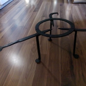 May include: A black, wrought iron trivet with a circular top, three legs, and a handle. The trivet is designed to hold hot pots and pans. The handle extends to the left, and the legs have rounded feet.