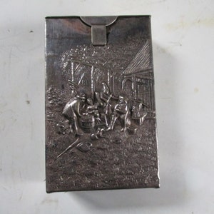 May include: Silver-colored rectangular box with a hinged lid. The front of the box features a detailed embossed scene of people gathered outdoors near a building. The scene includes figures, a barrel, and tools.