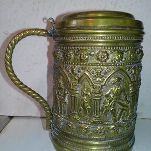 May include: An antique brass beer stein with a hinged lid and a twisted handle. The body is intricately decorated with relief carvings of figures and architectural elements. The stein has a tarnished, aged appearance.