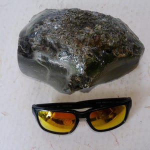 May include: A large, rough-textured, dark green rock sits above a pair of black sunglasses with yellow-orange lenses. The rock has a mottled appearance with areas of lighter green and brown. The sunglasses have a rectangular frame.