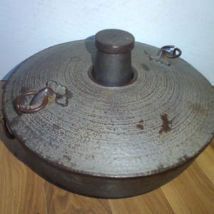 May include: A round, gray metal container with a lid and two metal handles. The container has a textured surface and a circular opening in the center.