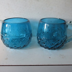 May include: Two blue glass mugs with a textured, diamond-shaped pattern. The mugs have a rounded shape and a handle.