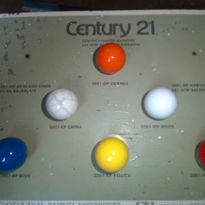 May include: A display of Century 21 cabinet knobs in various colors, including blue, white, yellow, orange, red, and a marbled gray. The knobs are labeled with their corresponding colors and product numbers.