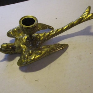 K&ouml;nnte beinhalten: Ein messingfarbener Vogel-Kerzenhalter. Der Kerzenhalter hat die Form eines Vogels mit ausgebreiteten Fl&uuml;geln und einem Schwanz. Der Kerzenhalter ist aus Messing gefertigt und hat eine goldene Farbe. Der Kerzenhalter ist f&uuml;r eine einzelne Kerze konzipiert.