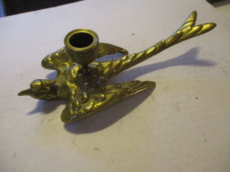 K&ouml;nnte beinhalten: Ein messingfarbener Vogel-Kerzenhalter. Der Kerzenhalter hat die Form eines Vogels mit ausgebreiteten Fl&uuml;geln und einem Schwanz. Der Kerzenhalter ist aus Messing gefertigt und hat eine goldene Farbe. Der Kerzenhalter ist f&uuml;r eine einzelne Kerze konzipiert.
