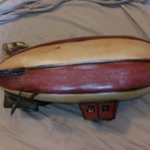 May include: A vintage-style blimp model, featuring a cream-colored top and a red lower section. The blimp has black accents, including a tail fin, propeller, and a small cabin. This decorative piece evokes a sense of nostalgia.