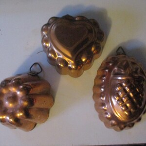 May include: Three vintage copper cake molds. One is heart-shaped, one is round with a fluted edge, and one is a flower shape.