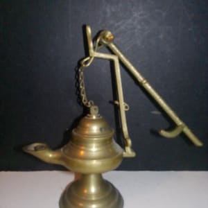 May include: A brass oil lamp with a decorative design and a hanging arm. The lamp has a round base and a spout for pouring oil.