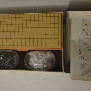 May include: A Go game set with a wooden board, black and white game pieces in clear containers, and a box with instructions. The board has a grid pattern with two black dots. The game pieces are in oval-shaped containers.