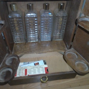 May include: A wooden bar cabinet with a hinged door and a pull-out drawer. The cabinet has four glass bottles with stoppers and a wooden rack with three cup holders.