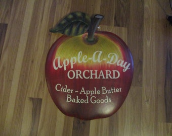May include: Wooden apple-shaped sign with the text "Apple-a-Day ORCHARD" in white script. The sign also lists "Cider - Apple Butter Baked Goods". The apple is red with a yellow center and a green leaf.