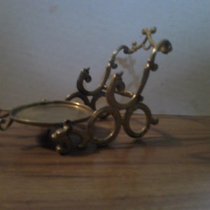 May include: A gold-colored metal stand with a round top and intricate scrollwork design. The stand is designed to hold a small object, such as a picture frame or a decorative item.