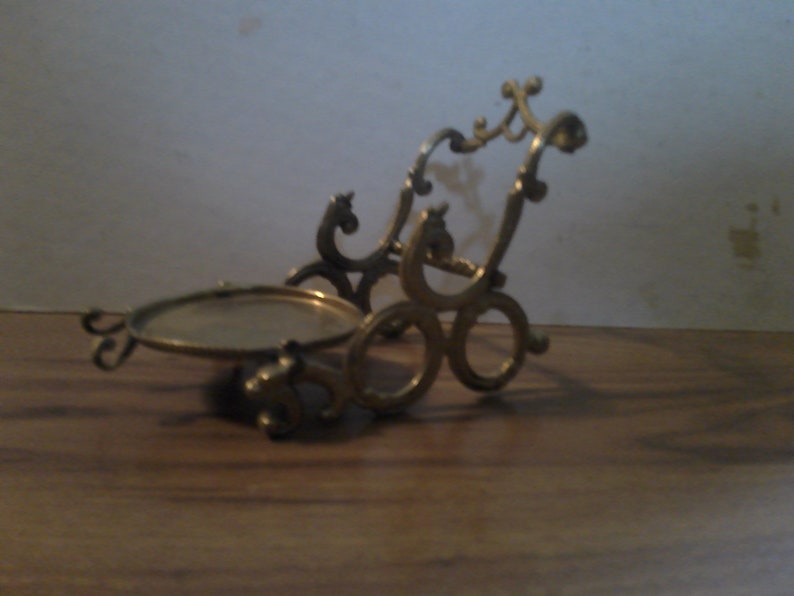 May include: A gold-colored metal stand with a round top and intricate scrollwork design. The stand is designed to hold a small object, such as a picture frame or a decorative item.