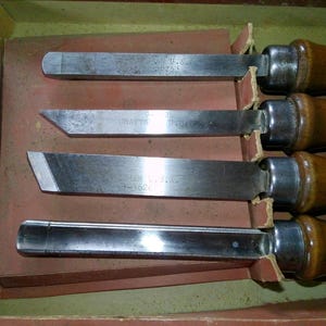 May include: A set of four woodturning chisels with polished metal blades and smooth wooden handles. Each blade features engraved text including the word Craftsman and U.S.A. The tools are arranged horizontally within a partitioned cardboard box.