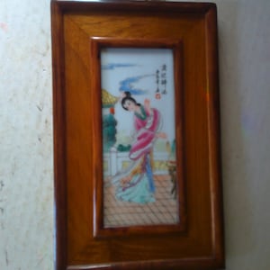 May include: A framed porcelain painting of a woman in a pink and green dress standing in a garden setting. The painting is surrounded by a wooden frame with a gold metal clasp at the top.