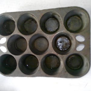 May include: A well-used, rectangular muffin tin with twelve circular molds. The metal pan has a dull, gray finish with some dark residue in the cups. Two small holes are on the side of the pan.