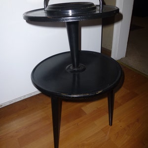May include: A vintage black side table with a unique design. It features a round top with a handle, a central support, and a lower round tier, all supported by three tapered legs. The table is made of wood and has a glossy finish.
